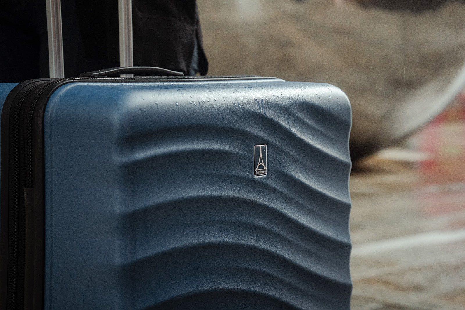 Close-up of a blue hard-shell carry-on suitcase with water-resistant exterior and textured wave design outdoors in rainy conditions.