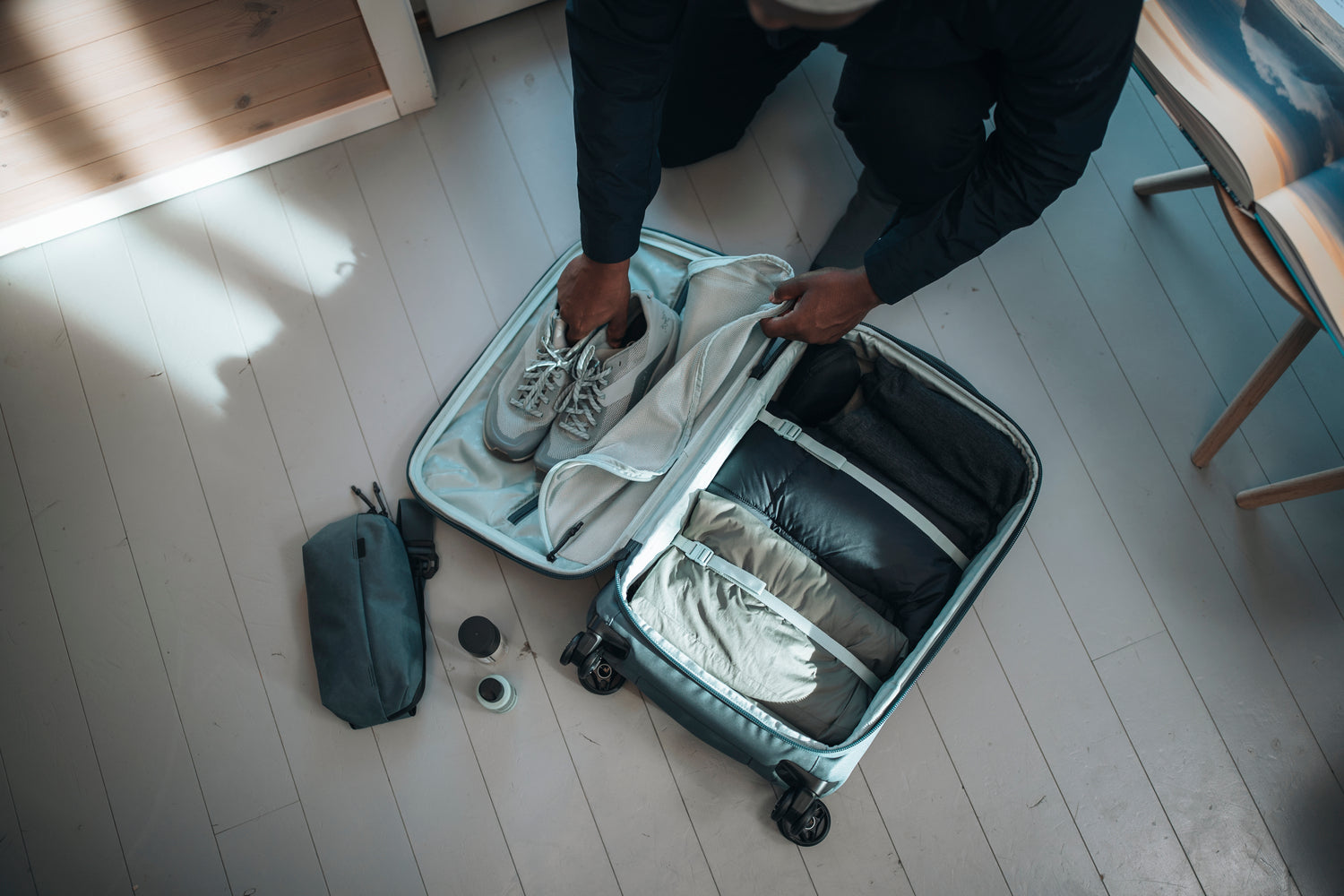 Traveler packing clothing and sneakers into an organized carry-on suitcase with divided compartments and travel essentials nearby.