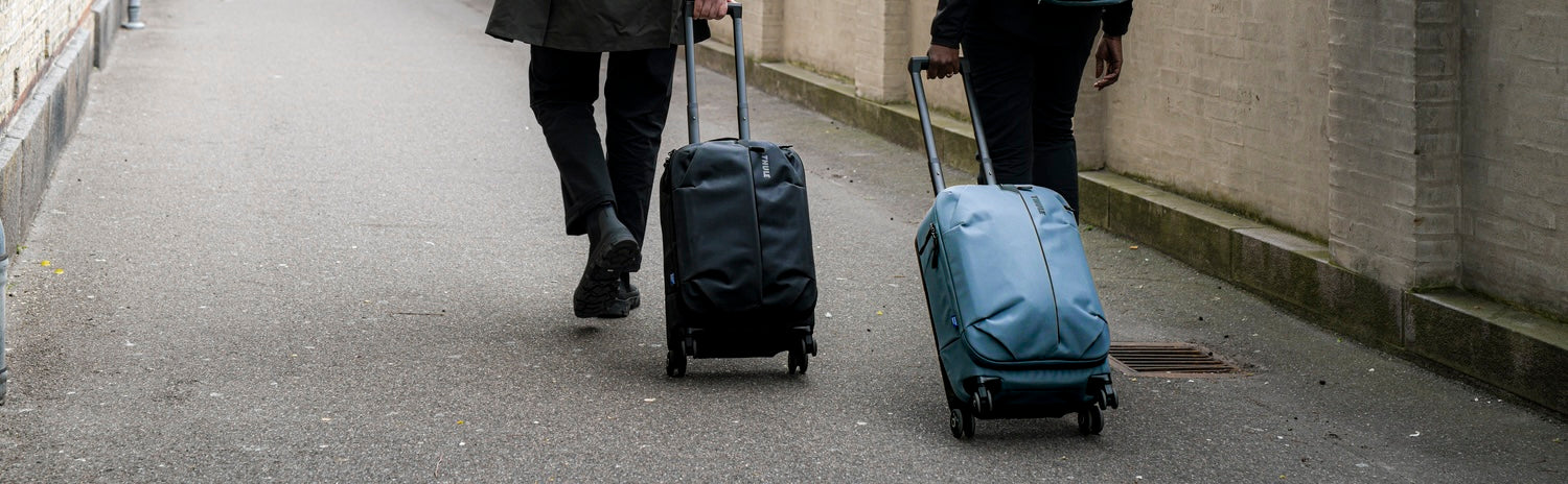 Two hikers carrying outdoor backpacks while trekking through a dense green forest in misty rainy conditions. Two travelers walking through a city street with wheeled carry-on luggage designed for urban and business travel.