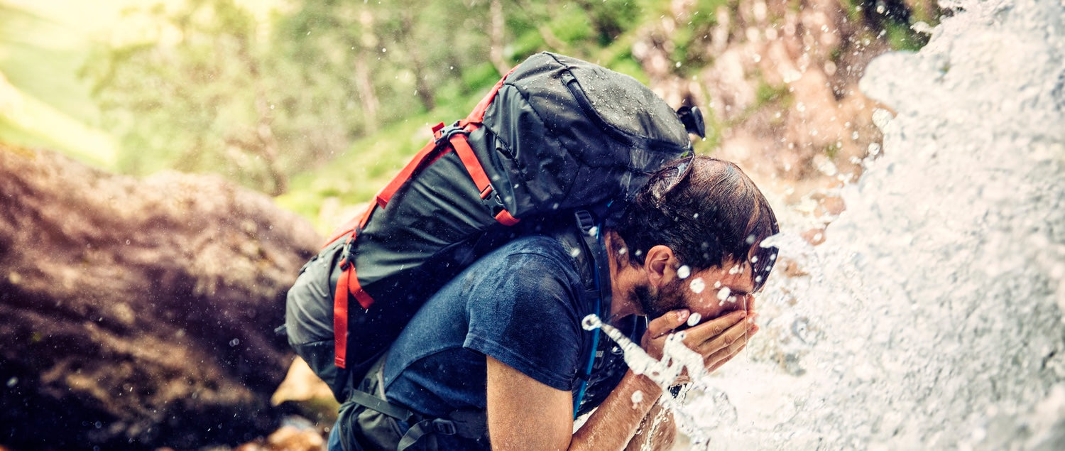 Hiker wearing a large outdoor backpack while drinking water beside a mountain waterfall during an adventure trek.