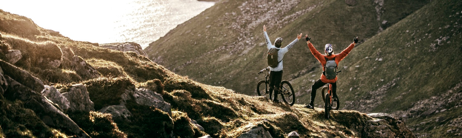 Two cyclists with outdoor backpacks celebrating on a mountain trail overlooking a scenic coastal landscape.