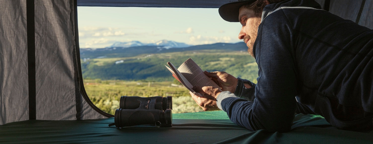 Traveler reading inside a rooftop tent with mountain views, outdoor gear, and binoculars during a camping adventure.