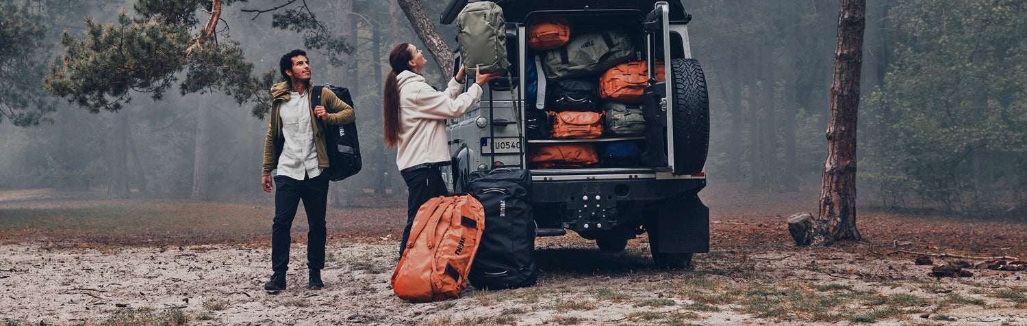 Two travelers unloading duffel bags and outdoor gear from an off-road vehicle during a forest camping adventure.
