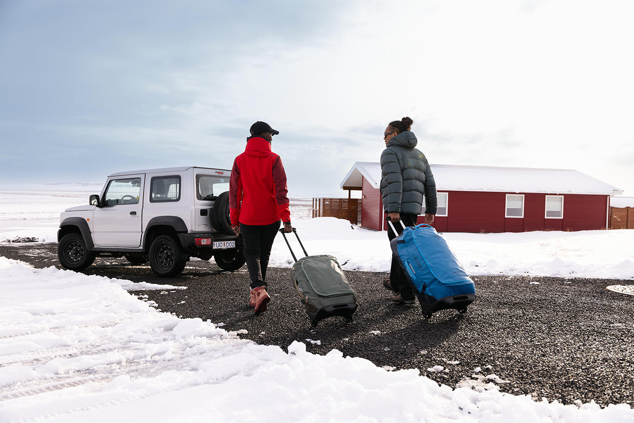 Two travelers walk across a snowy landscape pulling rolling duffel bags toward a white SUV parked beside a red cabin. One person wears a bright red jacket, while the other carries a blue bag and wears a gray puffer jacket. The scene feels cold, remote, and adventure-oriented.