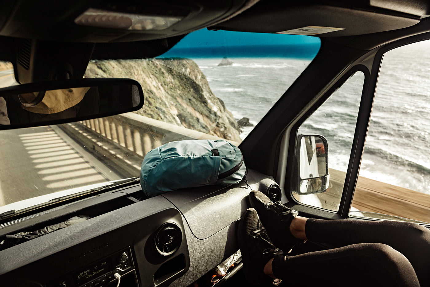Scenic coastal road trip view from inside a camper van, featuring ocean cliffs, winding highway views, and a travel backpack resting on the dashboard. Perfect for travel luggage, adventure gear, and outdoor lifestyle themes.