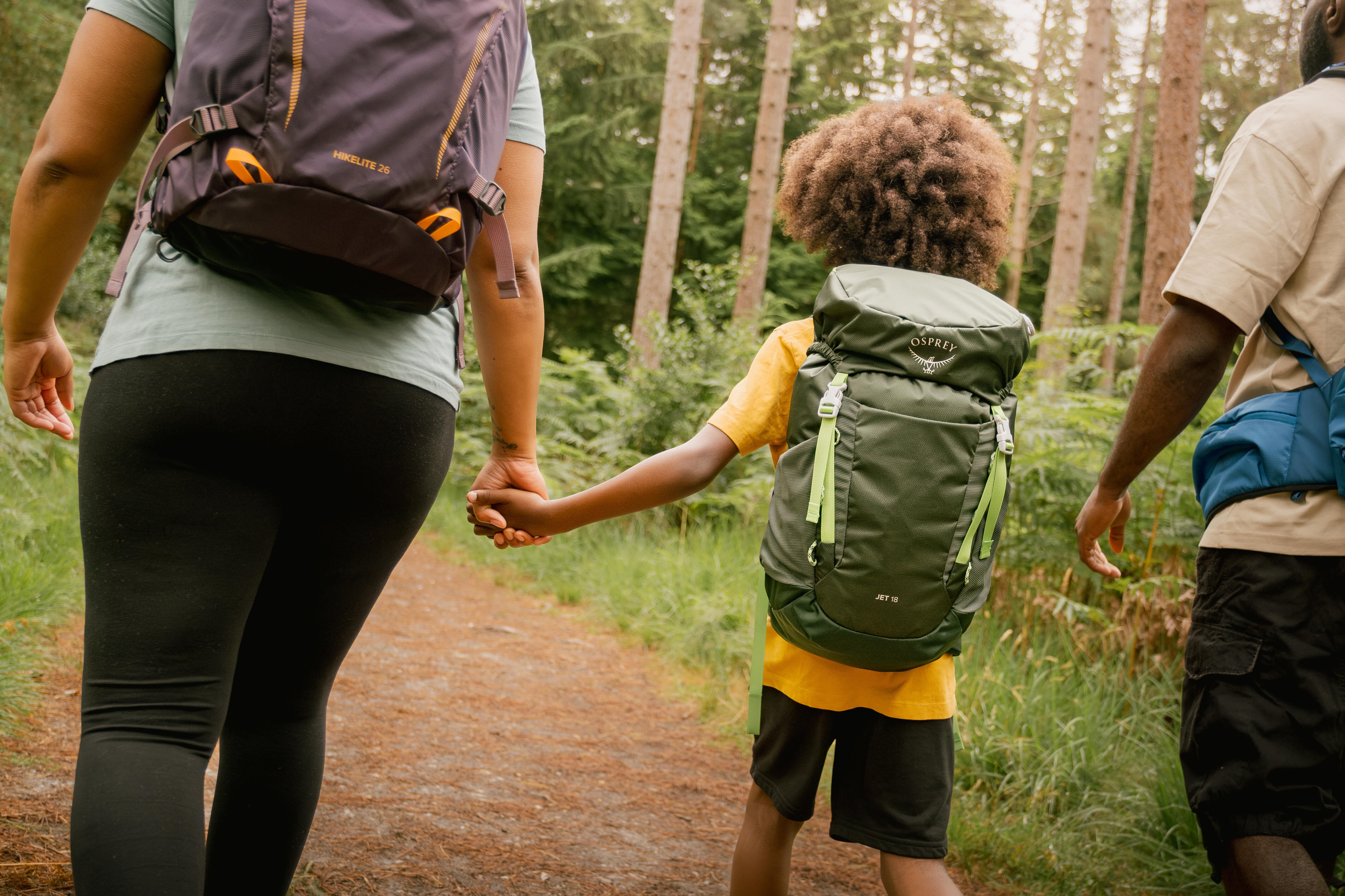 Two adults and a child walking on a trail in a forest, all wearing backpacks.