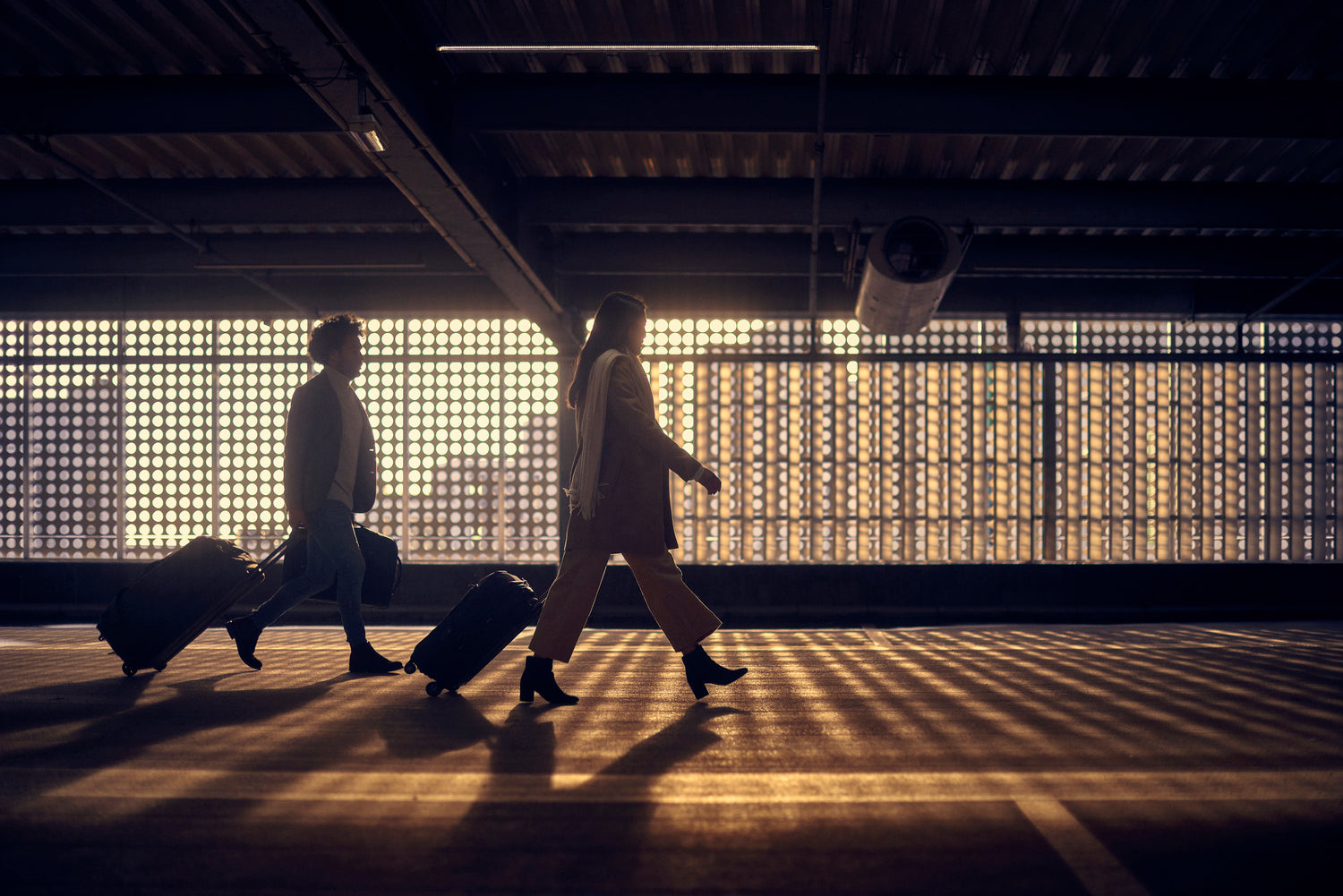 Two people with luggage walking through a train station with a patterned wall in the background.