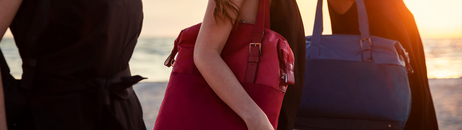 Three women walk along a beach at sunset carrying colorful travel bags. The center woman looks toward the camera with a bright red shoulder bag, while another carries a large blue tote. Warm golden light creates a stylish, relaxed travel atmosphere.