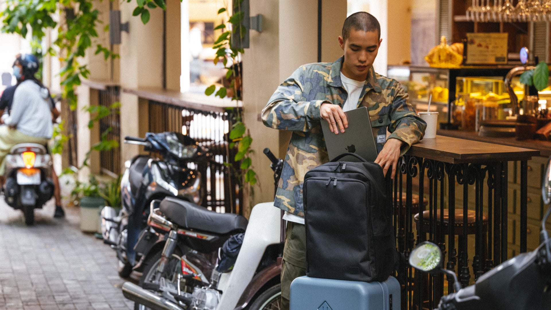 A man standing outside a café places a laptop into a black backpack resting on top of a suitcase. Motorbikes line the narrow street beside him, while warm café lighting and greenery create a relaxed urban travel atmosphere.