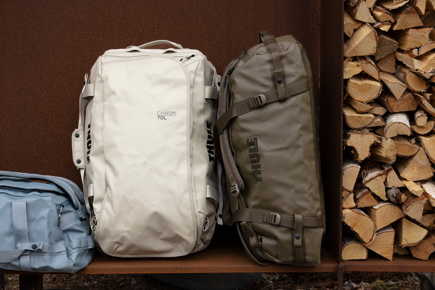 Three backpacks of different colors (white, beige, blue) on a shelf with stacked firewood in the background.