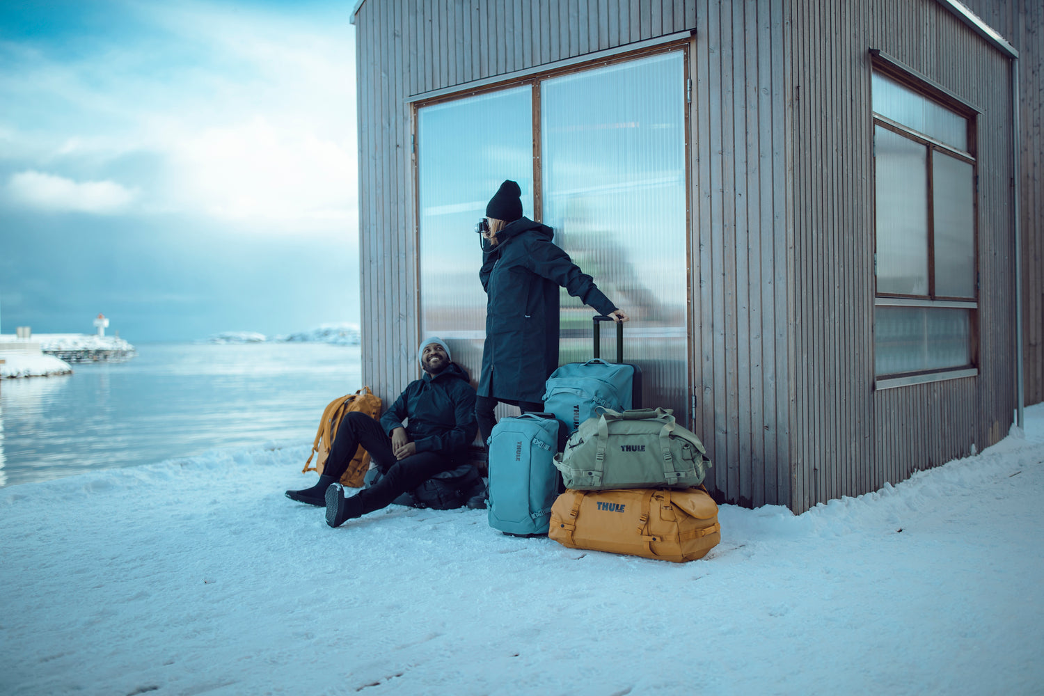 Two people with luggage by a building by a snowy landscape
