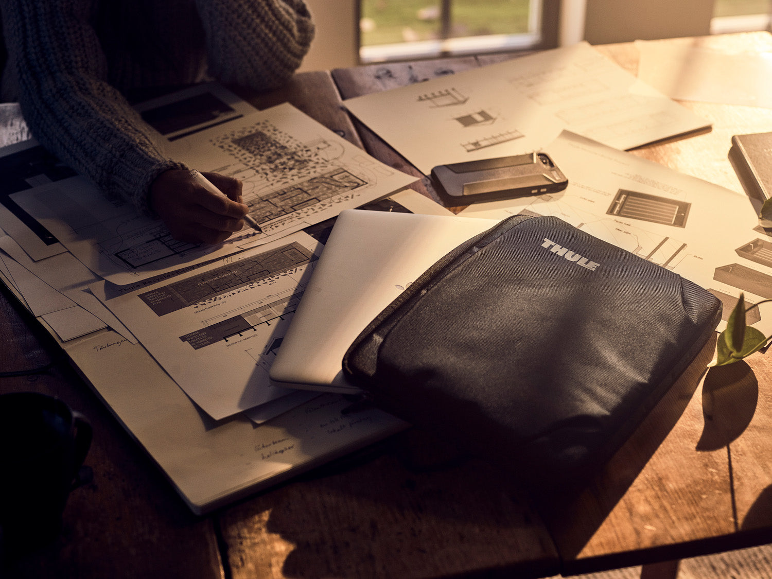 Person working at a desk with papers and a laptop, wearing a sweater.
