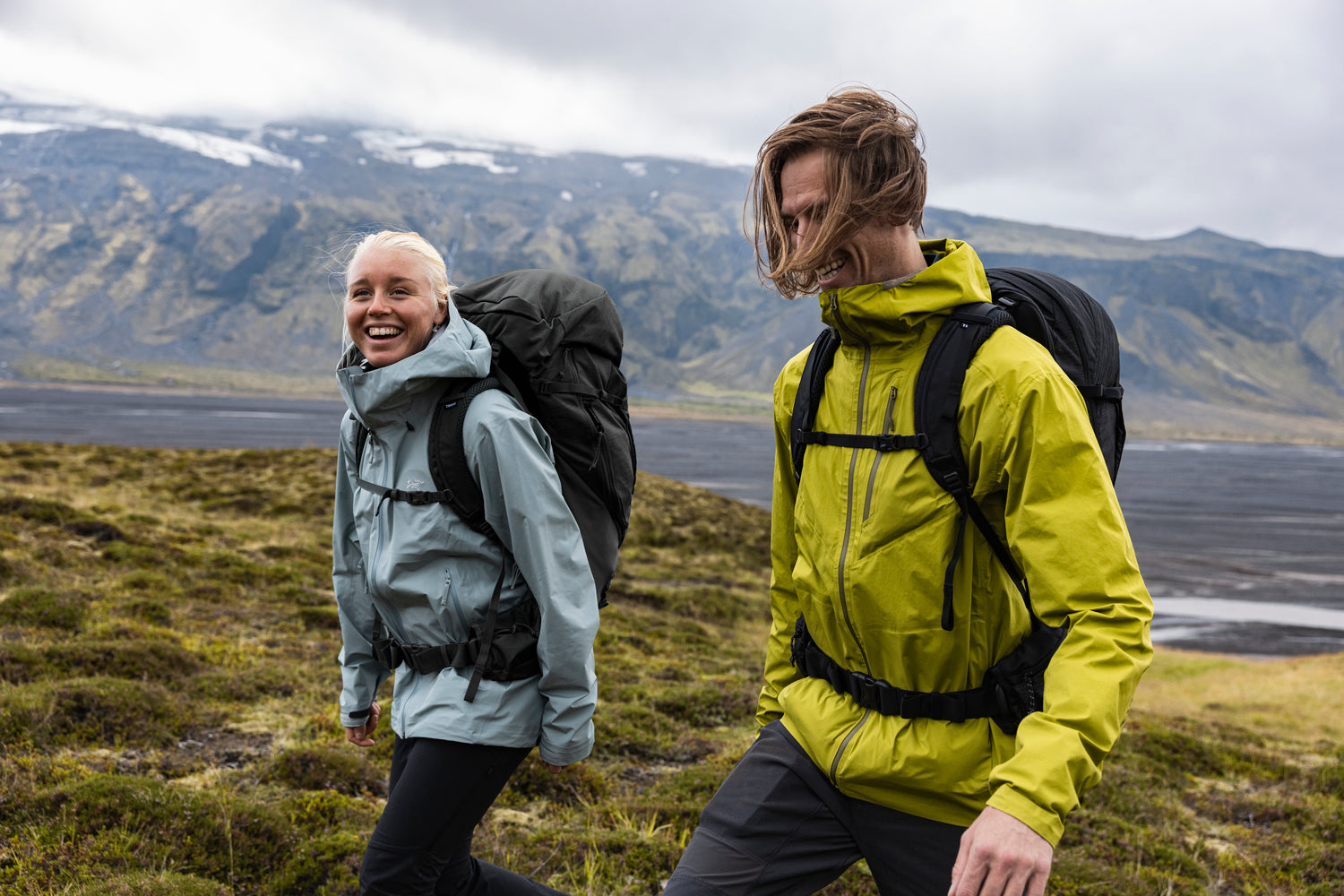 Two people hiking in a scenic outdoor setting with mountains and water in the background.